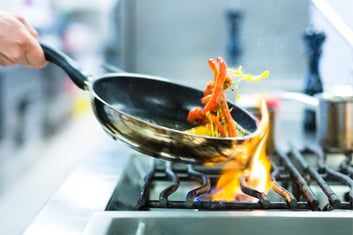 chef in restaurant kitchen at stove with pan doing flambe on food on a gas cooker